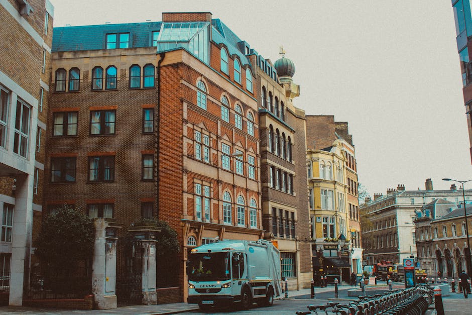 A street scene in an urban area featuring a collection of old and modern multi-story buildings with various architectural styles. The foreground shows a grey waste collection truck from Rubbish Removal Pimlico parked on the pavement, positioned near the entrance of a small gated area, with a black metal gate and stone pillars. The truck has a smooth, metallic finish, and the side panels reflect diffuse daylight. Surrounding the buildings are different facades, including red brickwork, ornate window frames, and decorative cornices, with some buildings topped by pitched roofs and domes. The street is lined with small bollards separating the sidewalk from the road, with bicycles secured to a bike rack on the right. The environment appears to be a busy city centre with light traffic and a generally overcast sky providing diffuse lighting, aligning with services like private rubbish collection and waste removal in the area, suggesting an active on-site clearance or alternative waste handling operation by Rubbish Removal Pimlico.