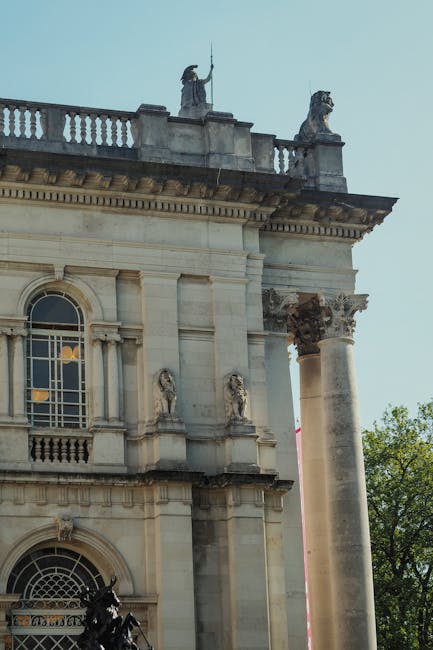 A close-up view of a historic building's upper façade, featuring classical architectural details such as tall, rounded windows with decorative frames, ornate stone carvings, and a solid stone cornice supported by Corinthian columns. The building's exterior is made of light-colored stone with visible textures and weathering. At the top, there are two stone sculptures of seated figures, each holding an object, positioned on the balustrade. The background shows a clear sky and green trees, indicating an outdoor setting. The scene is well-lit with natural daylight, highlighting the intricate stonework and architectural features, suggesting a location that may be associated with cultural or historic significance, potentially relevant to discussing private or alternative waste handling in the context of urban environments such as Pimlico, where rubbish removal services like Rubbish Removal Pimlico operate to assist with maintenance of historic areas.