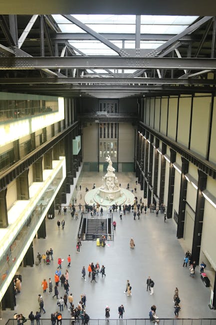 A street scene in an urban area featuring a collection of old and modern multi-story buildings with various architectural styles. The foreground shows a grey waste collection truck from Rubbish Removal Pimlico parked on the pavement, positioned near the entrance of a small gated area, with a black metal gate and stone pillars. The truck has a smooth, metallic finish, and the side panels reflect diffuse daylight. Surrounding the buildings are different facades, including red brickwork, ornate window frames, and decorative cornices, with some buildings topped by pitched roofs and domes. The street is lined with small bollards separating the sidewalk from the road, with bicycles secured to a bike rack on the right. The environment appears to be a busy city centre with light traffic and a generally overcast sky providing diffuse lighting, aligning with services like private rubbish collection and waste removal in the area, suggesting an active on-site clearance or alternative waste handling operation by Rubbish Removal Pimlico.